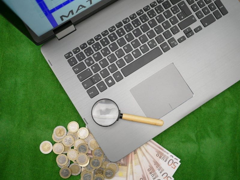 Ein Laptop liegt auf mehreren Geldscheinen, daneben liegen Münzen. Auf dem Laptop liegt eine Lupe.