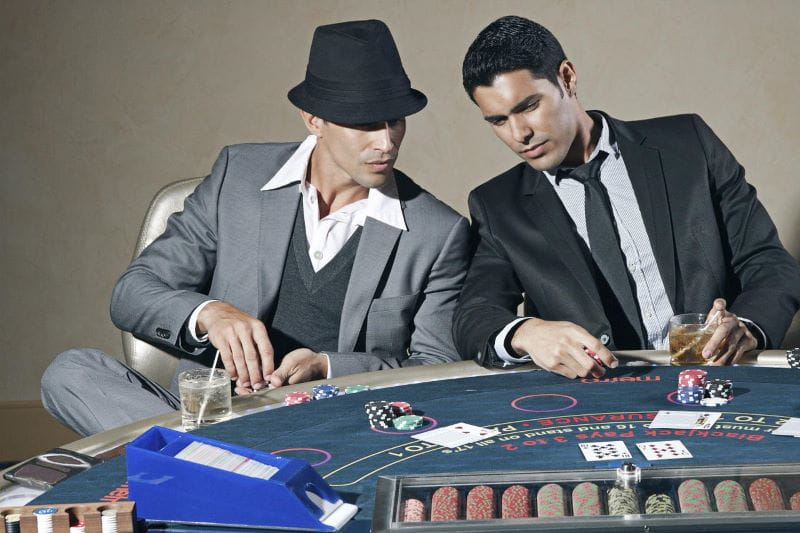 Zwei elegant gekleidete Männer sitzen an einem Pokertisch, auf dem Karten, Spielchips und Gläser stehen.